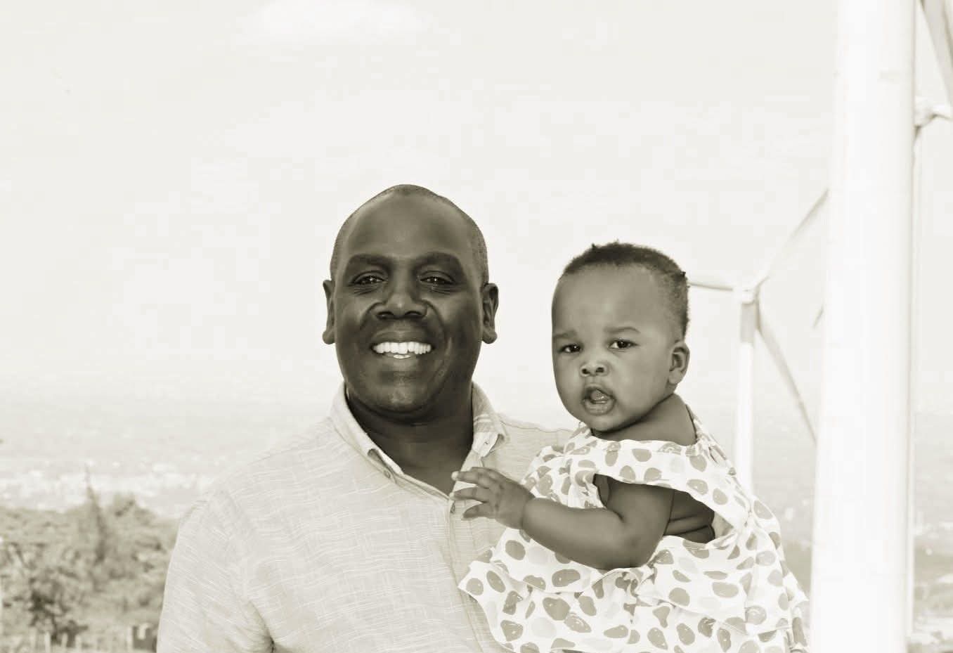 Sepia portrait of Enock Change holding a child with a wind turbine behind